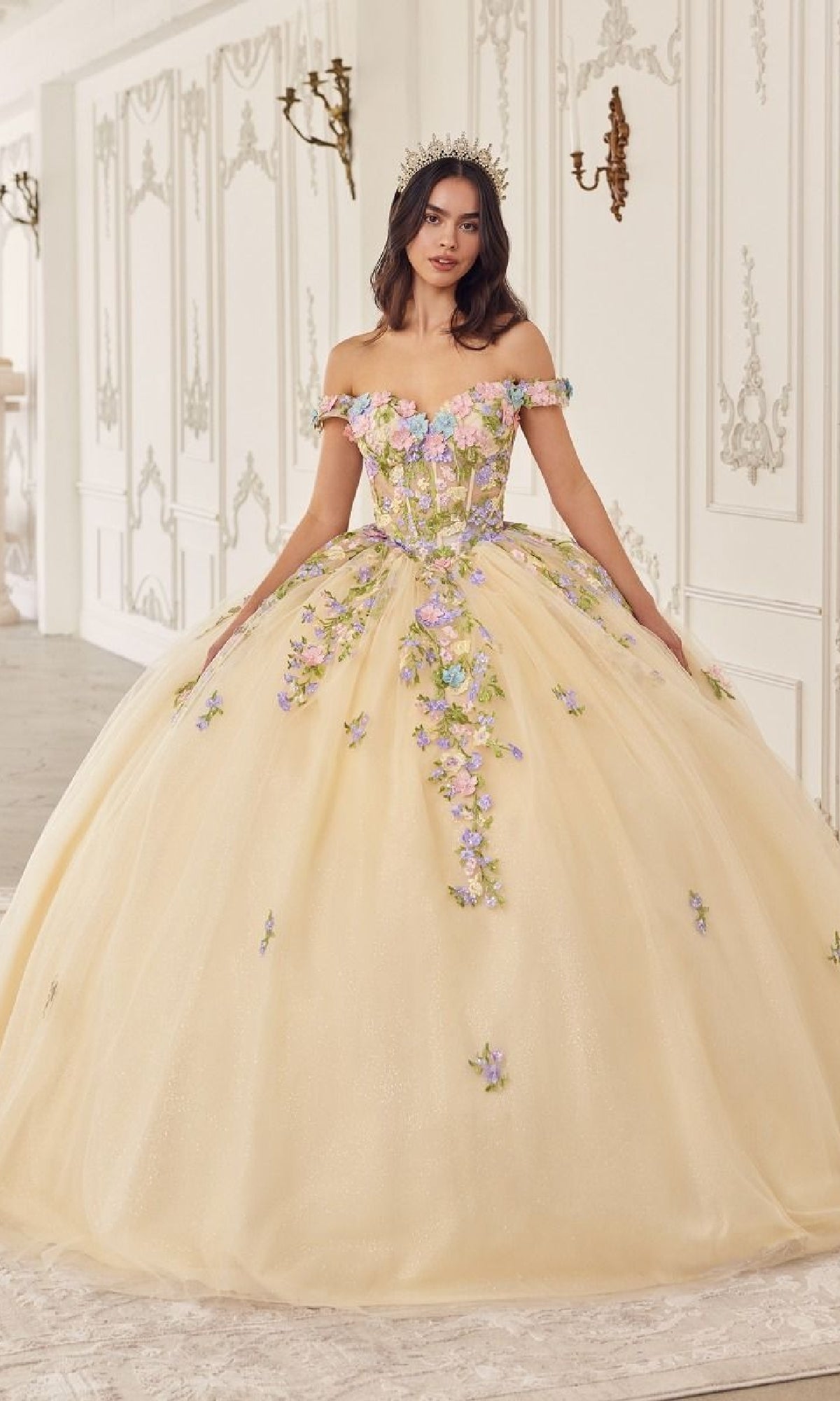 A woman in the Ladivine Quinceanera Dress 15724—an off-the-shoulder yellow ball gown with pastel floral appliqué and a tiara—stands gracefully in an ornate white room.